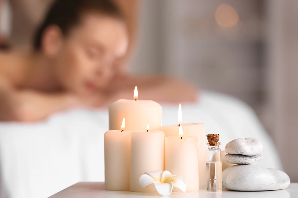 Candle-lit spa setting with lit pillar candles, a frangipani flower, essential oil and stacked spa stones, a woman relaxing on a massage table behind