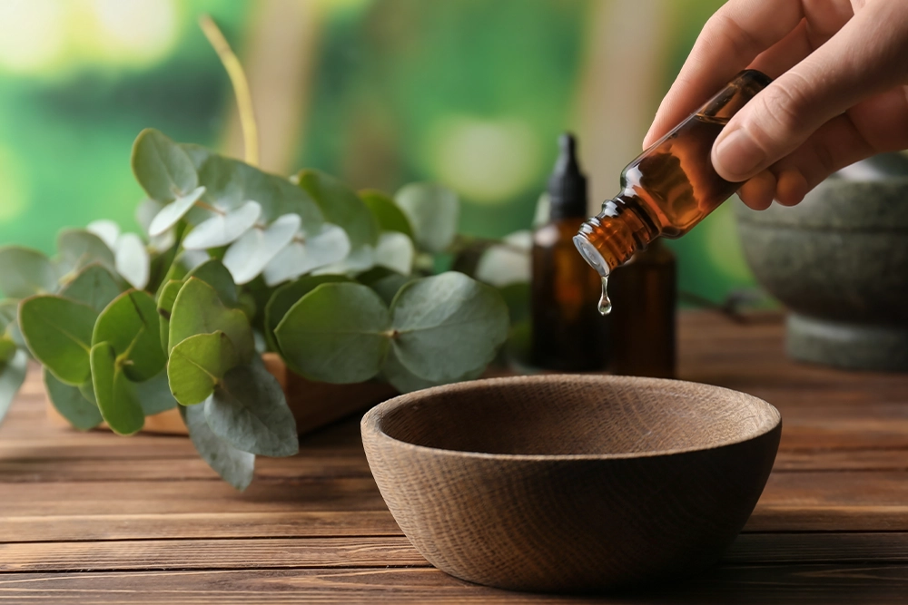 Natural essential oil being dripped from an amber dropper bottle into a wooden bowl beside fresh eucalyptus